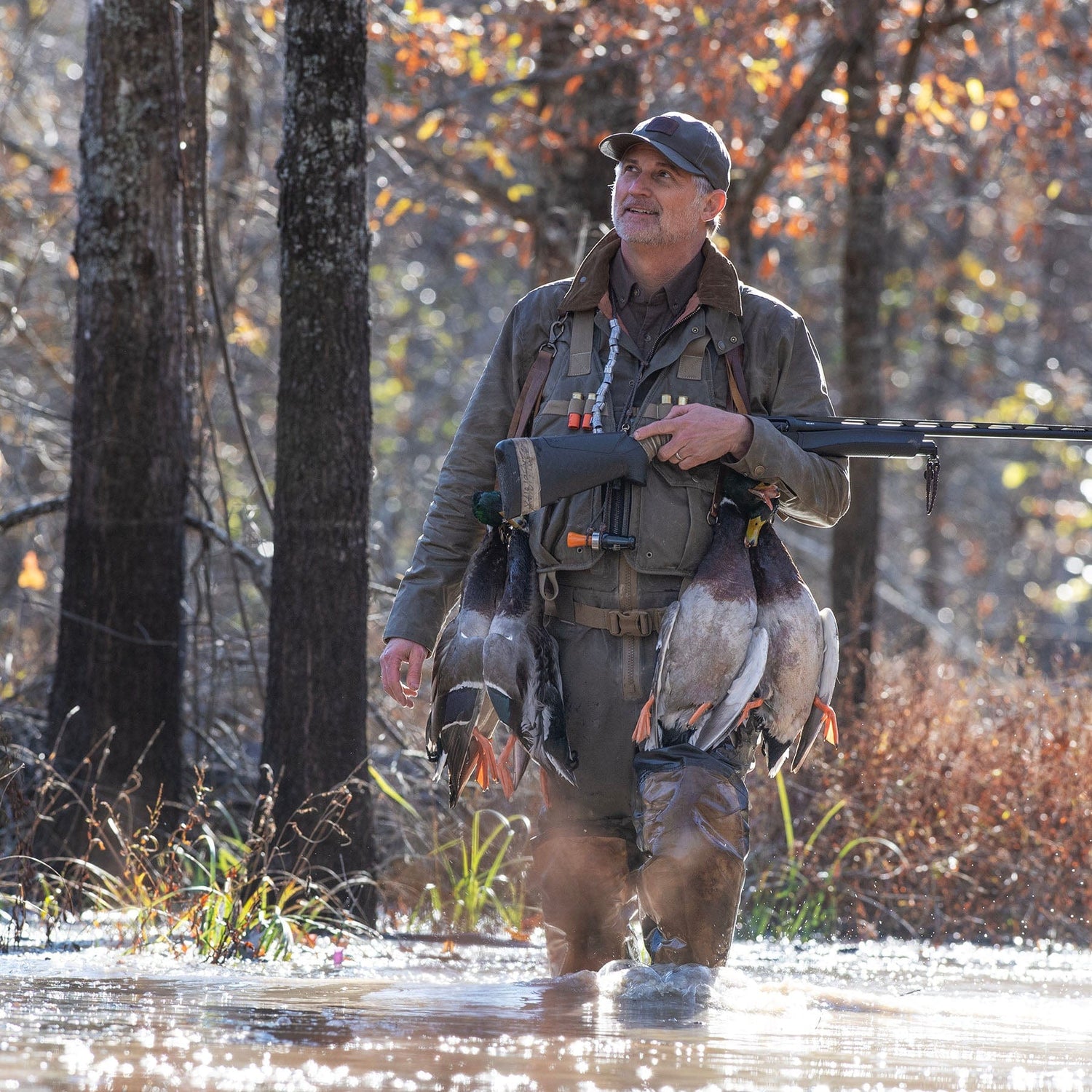 A man in outdoor gear stands in shallow water holding a rifle and several ducks, surrounded by trees in autumn.