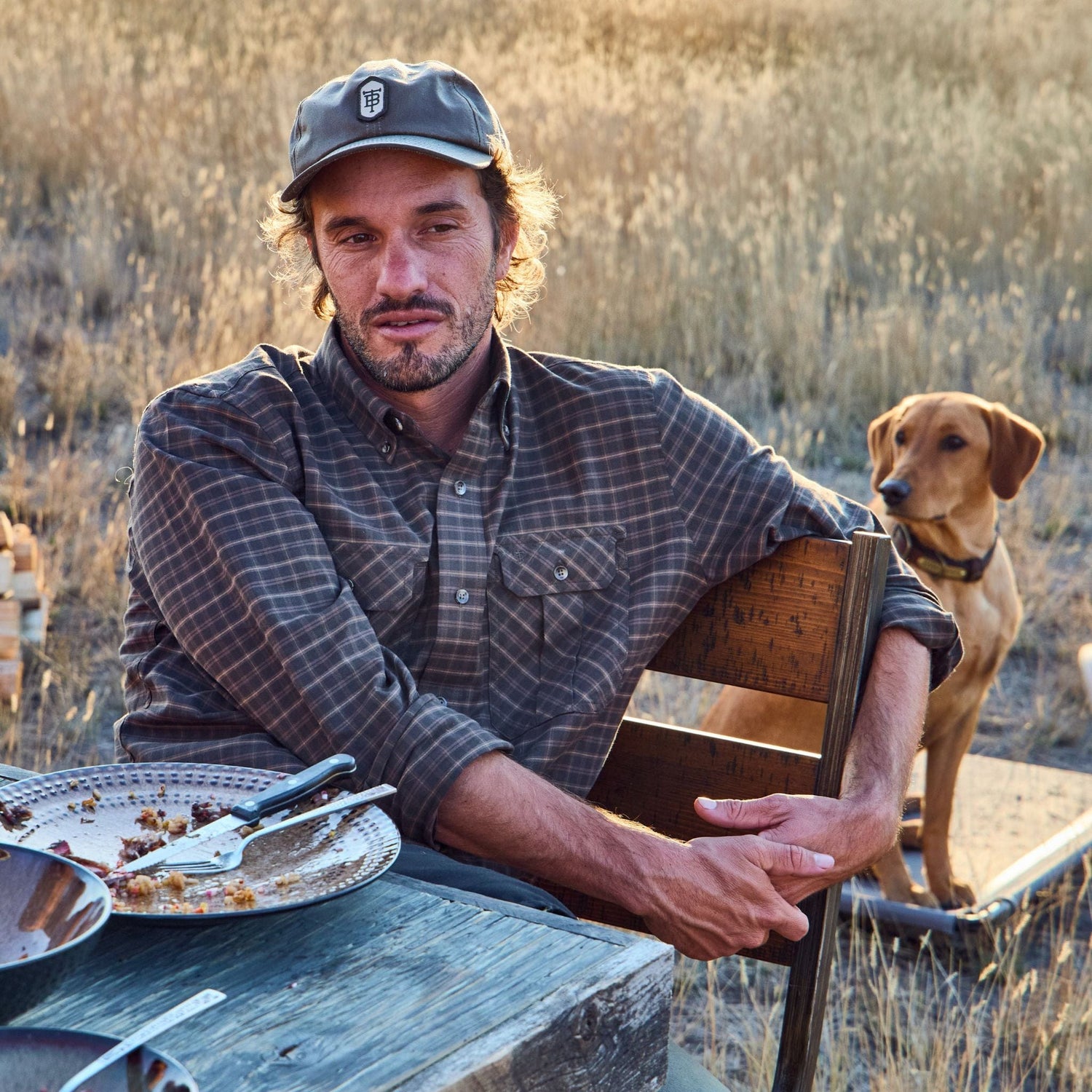 Man in a plaid shirt and cap sits outdoors at a table with empty dishes; a dog stands nearby in grassy field.