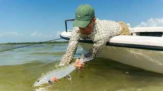 A person on a boat releases a large fish back into shallow, clear water under a blue sky.
