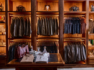 Wooden shelves filled with neatly arranged jackets, shirts, and jeans; a central table displays folded shirts, glass decanters, and leather bags.
