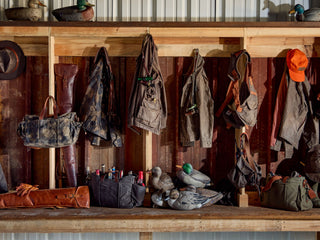 Wooden rack holding hunting gear, hats, coats, bags, decoy ducks, and drinks against a corrugated metal wall in a rustic setting.
