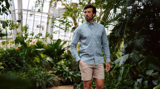 A man in a blue short-sleeve button-up shirt and tan shorts stands near a body of water with trees in the background.