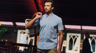 A man wearing a brown plaid shirt and khaki pants stands outdoors in front of trees with autumn leaves, smiling and looking to the side.