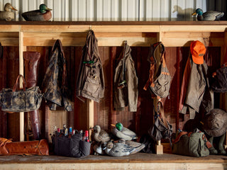 Wooden shelf with hanging outdoor gear, bags, jackets, an orange cap, duck decoys, and assorted hunting supplies arranged on and below the shelf.