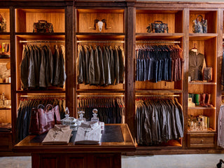 A wooden clothing display with jackets, shirts, and pants hanging neatly, shelves with bags, boots, and accessories, and a table with folded shirts and glassware in the foreground.