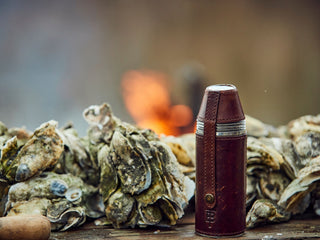 A brown leather-wrapped thermos stands on a wooden surface beside a pile of oyster shells, with a fire burning in the blurred background.