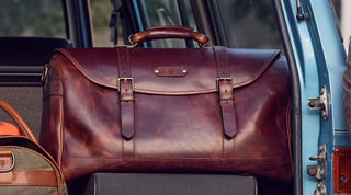 A large brown leather duffel bag with buckles is placed in the open trunk of a blue vehicle.