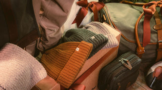 Knit beanies in brown, green, and beige are displayed on cardboard boxes, surrounded by travel bags and packages with brown ribbons.