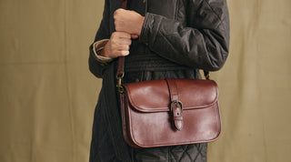A person wearing a quilted jacket carries a brown leather crossbody bag with a buckle closure.