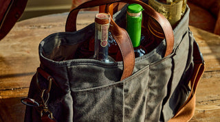 A person places a bottle of whiskey into a black duffel bag already containing other bottles, while carrying a brown shoulder bag outdoors.