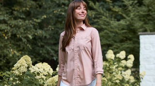 A woman stands outdoors by a wooden structure, holding a white mug and wearing a cream-colored button-up shirt and jeans. Trees are visible in the background.