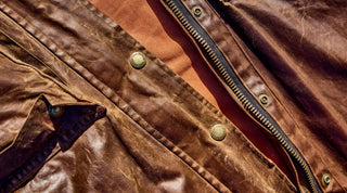 Close-up of a brown leather jacket with a visible zipper and metal snap buttons, worn over a reddish-brown shirt.