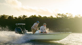 A man in a plaid shirt and cap is fly fishing outdoors, seen from behind, casting a fishing line against a partly cloudy sky.