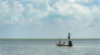 Three men outdoors; one in front holding a fishing rod, another behind standing and pointing, and a third sitting, all wearing casual shirts and caps with trees in the background.