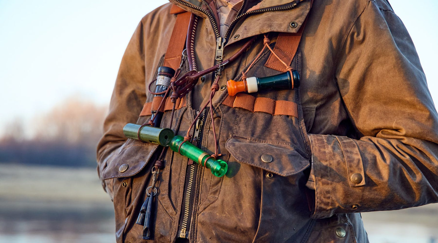 Person wearing a brown jacket and chest harness with multiple duck calls attached, standing outdoors near a body of water.