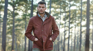 A man in a brown jacket with hands in his pockets stands outdoors in a forest with tall trees in the background.