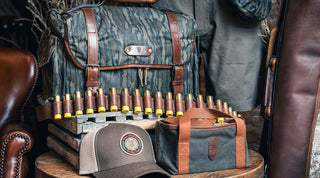 A camouflage hunting bag, shotgun shells lined up on a wooden crate, a brown and beige cap, and a small canvas bag are displayed together indoors.