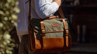 A person wearing a light-colored shirt carries a green and brown leather messenger bag with straps over their shoulder.