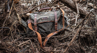 A green and brown camouflage duffel bag with leather straps hangs outdoors among dried brown foliage.