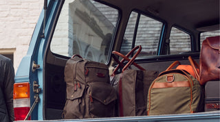 Open trunk of a blue vehicle loaded with various bags, including backpacks and duffel bags, parked near a light-colored building.