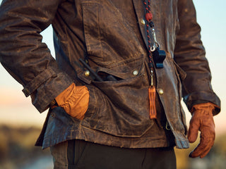Person wearing a brown waxed jacket and orange gloves, with tools and whistles hanging from their neck and a pair of glasses partially visible in a pocket.
