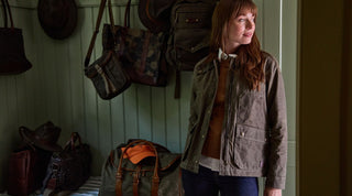 A woman wearing a brown jacket stands in a room with several bags and hats hanging on the wall and a duffel bag on the bed.