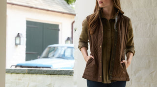 A woman stands on a porch wearing a brown quilted vest over an olive shirt with her hands in her pockets. A vintage blue car is parked outside in the background.