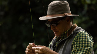 A person wearing a brown hat and jacket stands in tall dry grass holding a shotgun, surrounded by leafless trees and brush.