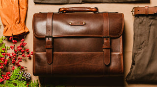 A brown leather briefcase with TOM embossed on a metal nameplate, surrounded by leather gloves, a belt, trousers, and holiday greenery.