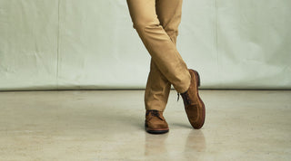 Person standing on stairs holding a black backpack in one hand, wearing olive pants and brown shoes, with only the lower body visible.