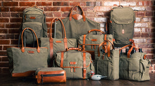 A collection of green canvas and brown leather bags and totes of various sizes are arranged in front of a brick wall.