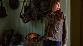 A woman in casual outdoor clothing stands in a doorway with hands in pockets; behind her are backpacks, hats, and bags hanging on wall hooks.