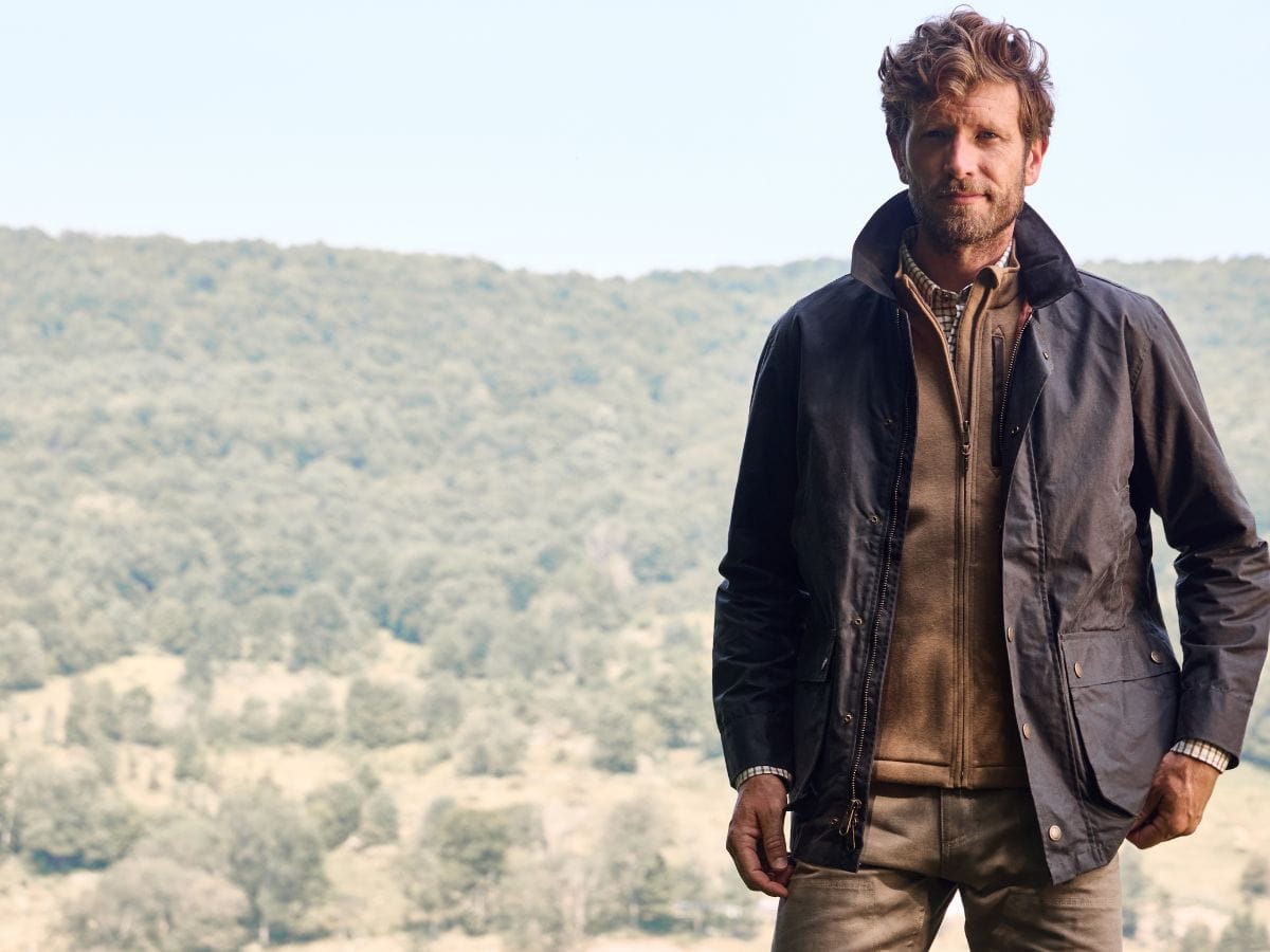 Man standing outdoors in front of a hilly landscape, wearing a dark jacket and light-colored clothing.