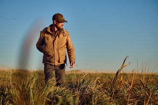 A person in a tan jacket and cap walks through a grassy field under a clear blue sky.