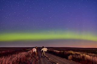Two people stand on a rural road under a starry sky with green aurora borealis in the background.