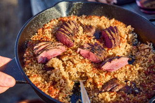 A skillet of cooked rice with grilled, sliced meat on top, served outdoors with a spoon.