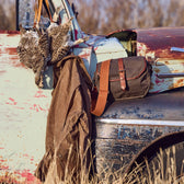 A rusted vintage truck with pheasants and a Tom Beckbe Canvas Blind Bag on the door, set against grass and trees.