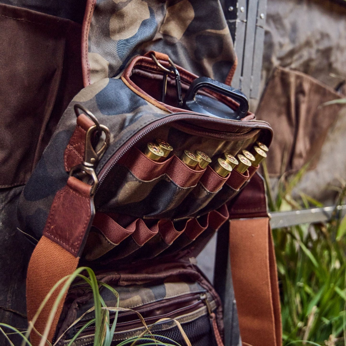 Open Tom Beckbes Classic Camo Blind Bag with double-shell pockets, revealing shotgun shells on grass and a dangling strap.
