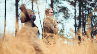 Two people in brown jackets stand in tall grass with shotguns, surrounded by trees in an outdoor setting.