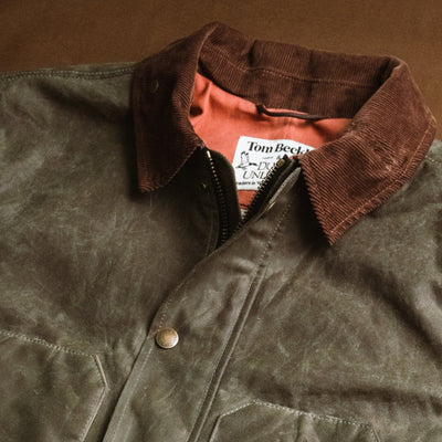 A waxed canvas jacket with a brown corduroy collar and a visible label on a brown background.