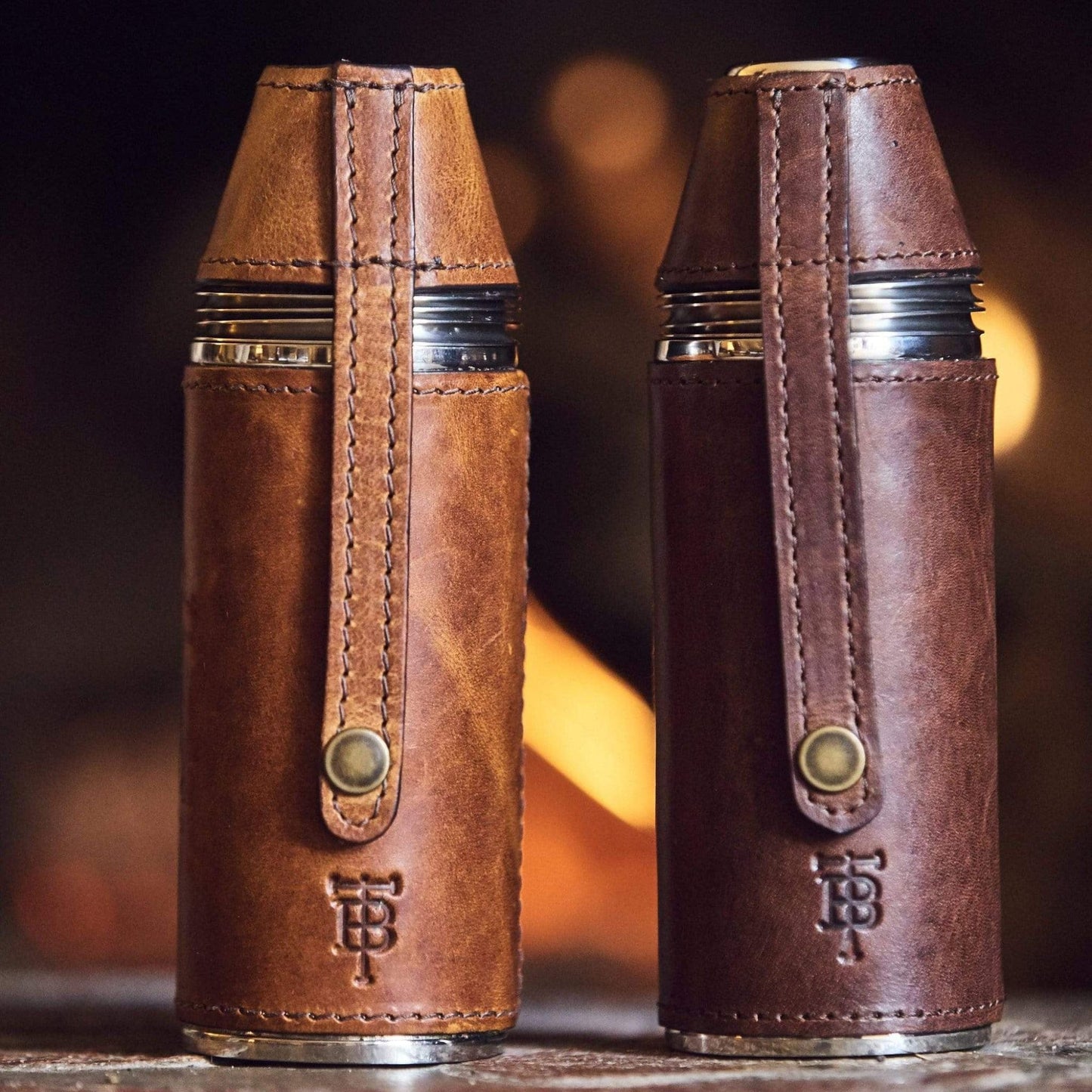 Two bottle flasks wrapped in full-grain leather embossed with TB logo sit on a table by a fire.