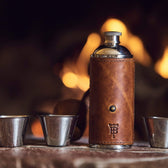 Bottle flask in chestnut leather with embossed TB logo and 3 toasting cups sit on a table by a fire.