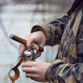 Man wearing a camo waxed jacket and cigar pours bourbon from a bottle flask into a stainless cup.