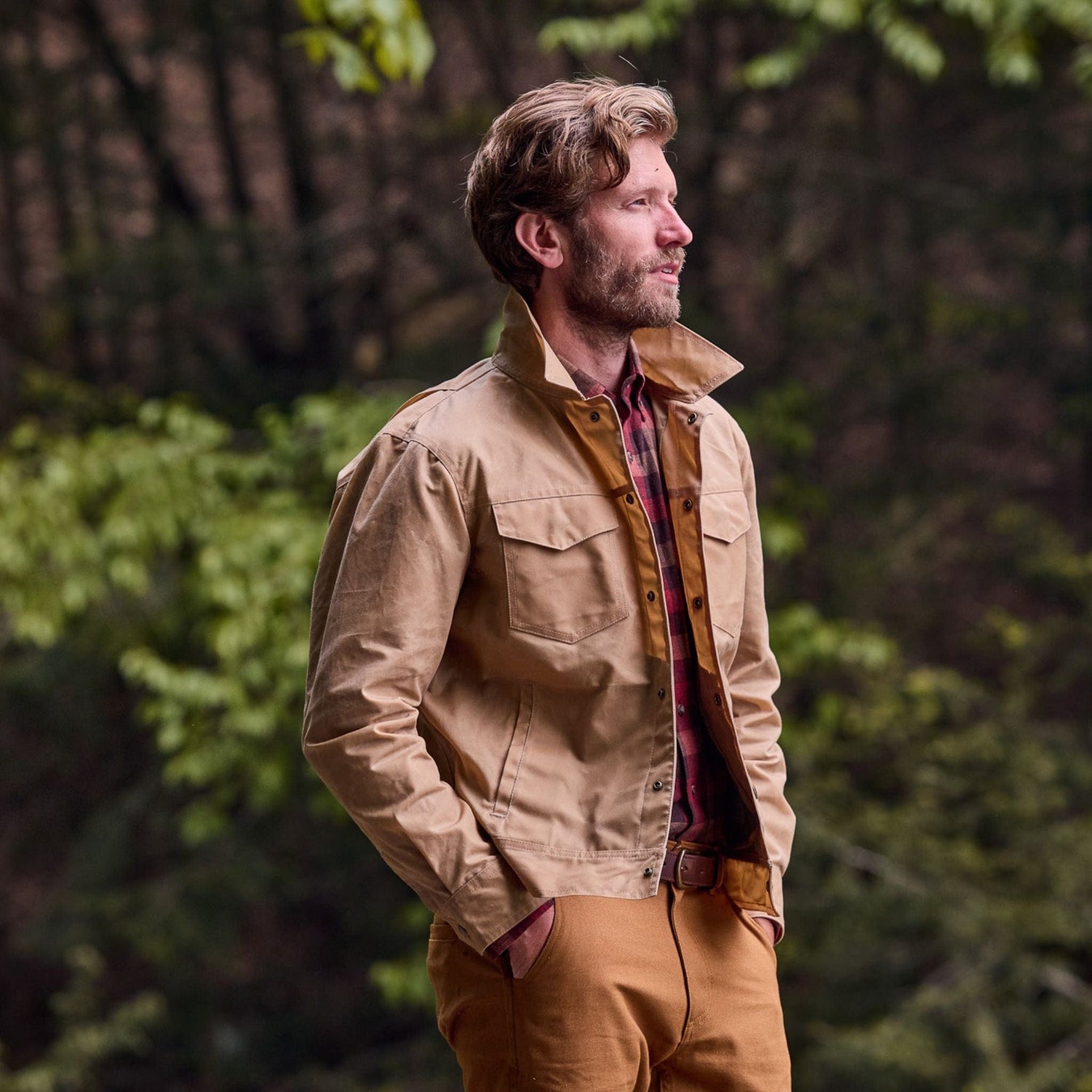 A man in a Tom Beckbe Chatom Jacket and brown pants stands with hands in pockets, against green foliage.