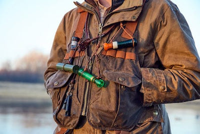 A person wearing a brown jacket and vest with multiple duck calls and accessories near a body of water.