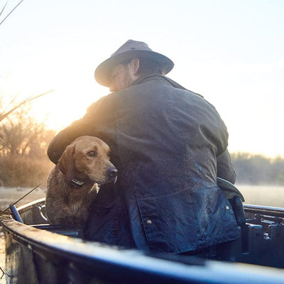 Man in a leather jacket and hat sits in a boat with a dog, looking towards the water on a misty morning.