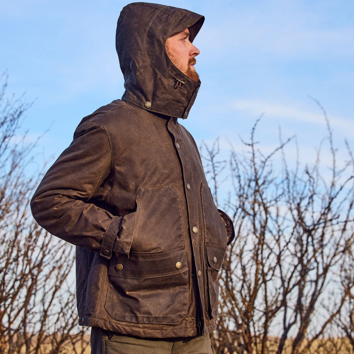 Someone wearing a hooded Fowler Jacket by Tom Beckbe stands outside with hands in pockets, against leafless shrubs.