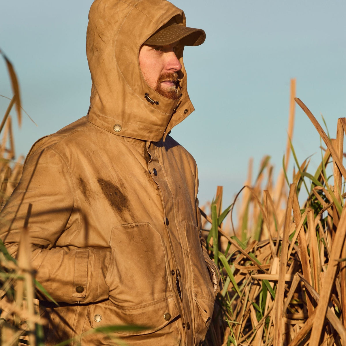 A person in a Tom Beckbe Fowler Jacket and cap stands in tall grass, gazing into the distance.