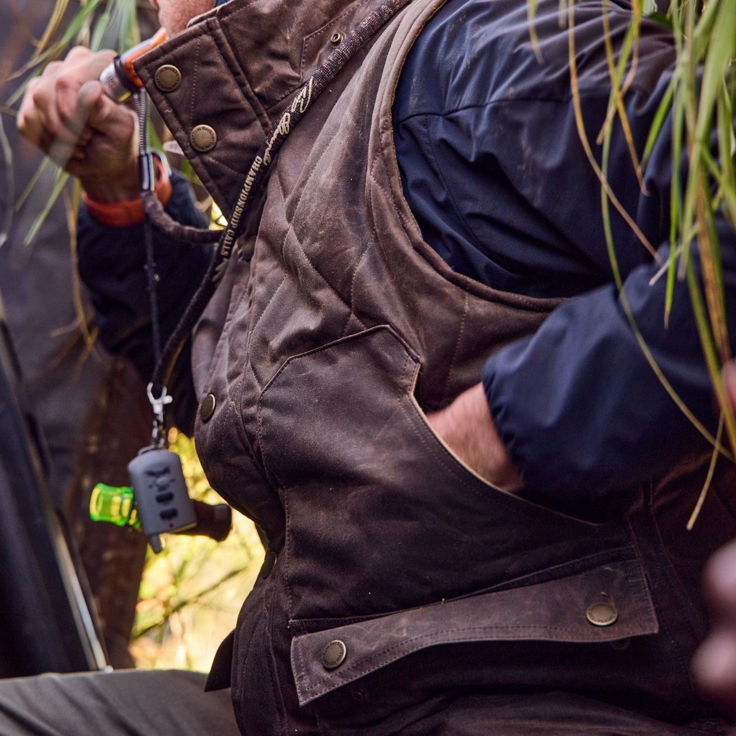 Person in a Tom Beckbe Fowler Vest with a whistle, seated outdoors against a lush foliage backdrop.