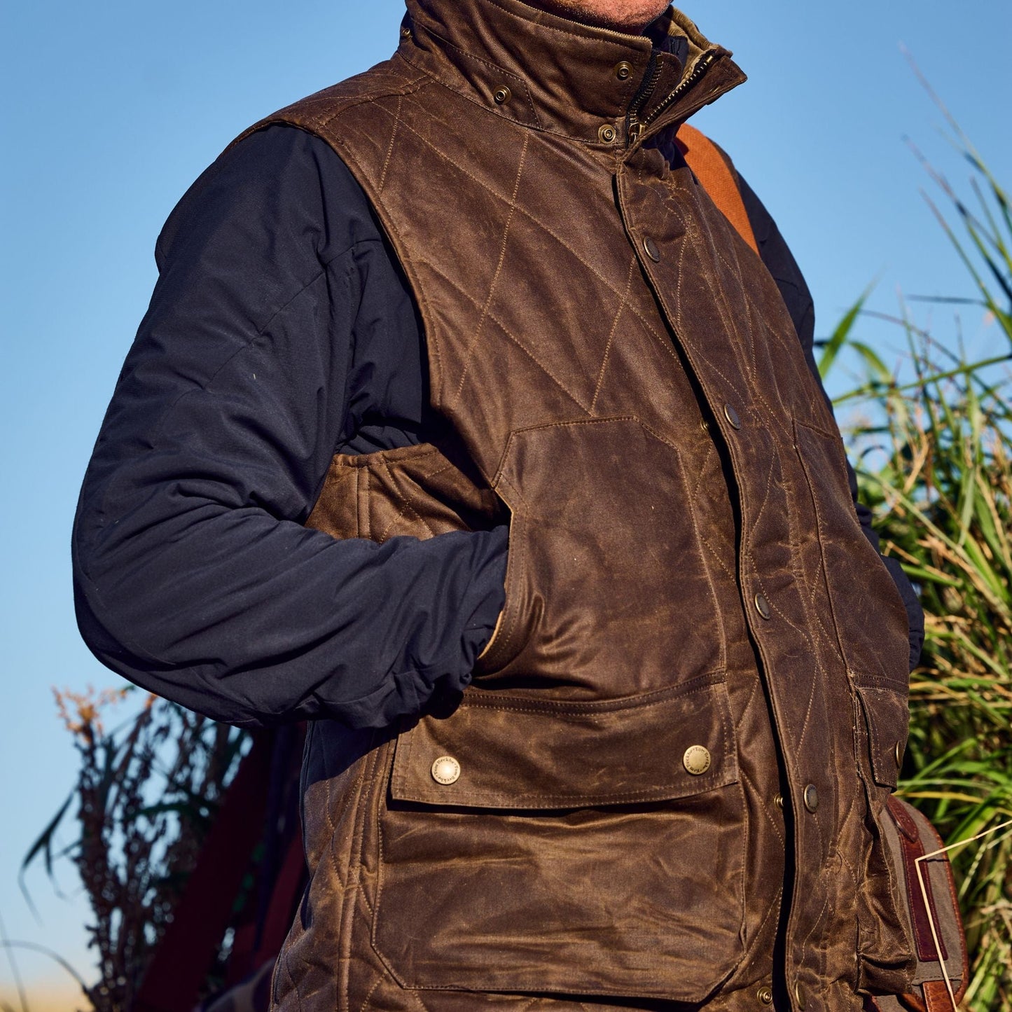 Someone outdoors in the Tom Beckbe Fowler Vest and a dark shirt, hands in pockets, with plants behind them.